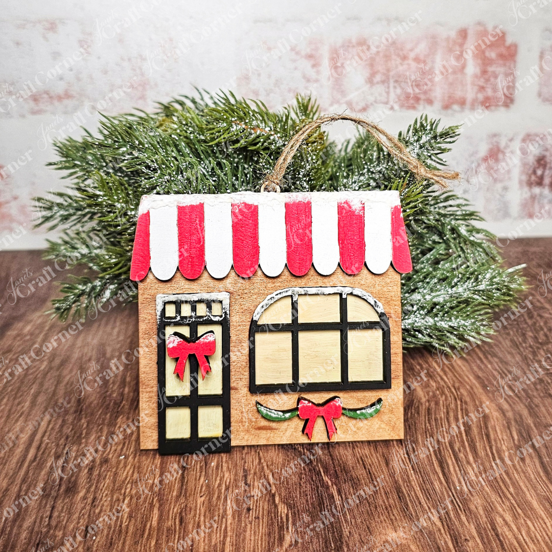 A quaint, decorative wooden house with a red and white striped roof from Janet's Craft Corner features a black door and windows with red bows. It's included in the Holiday Village Ornament Set - Early Access, charmingly placed among green pine branches on a wooden surface.