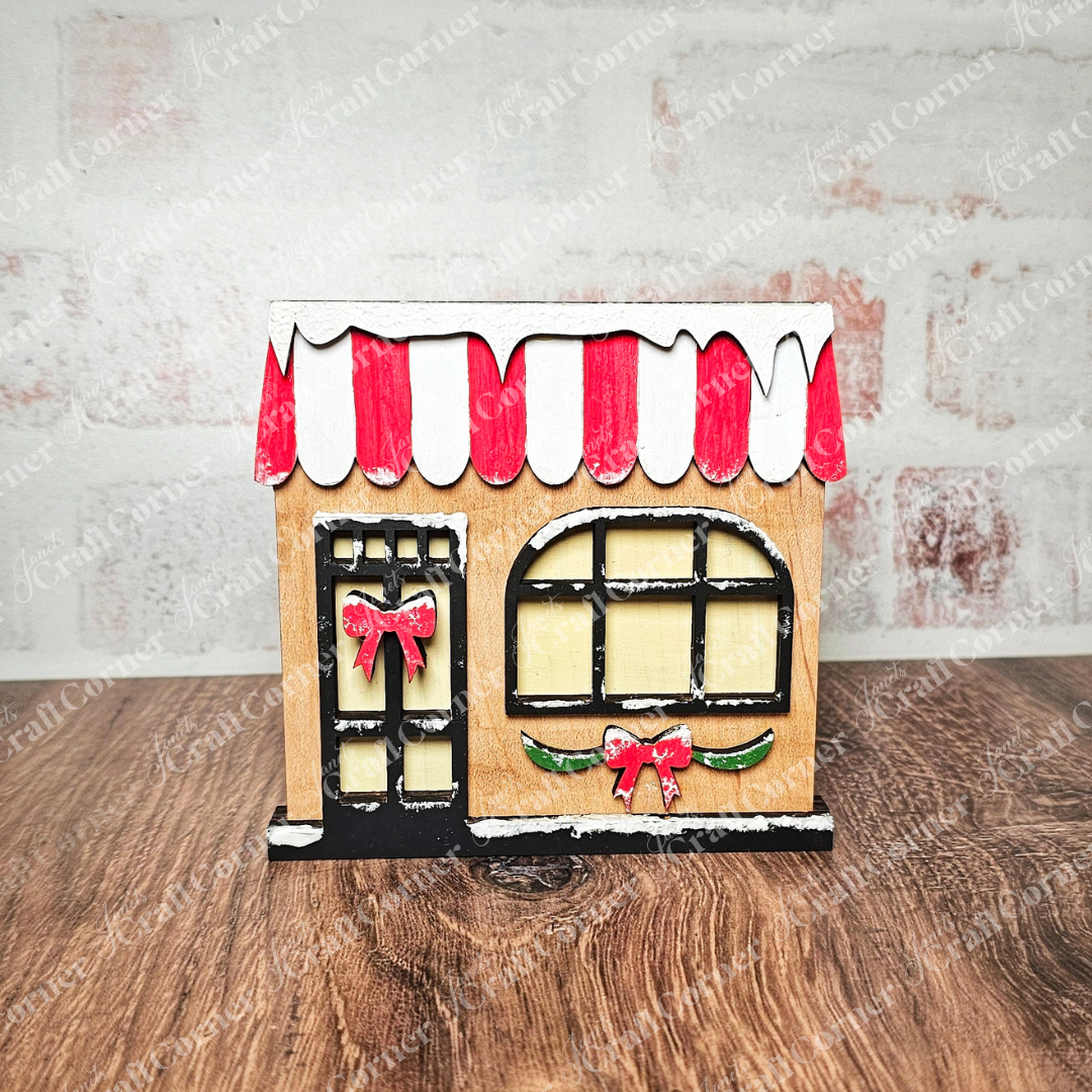 This delightful wooden miniature house from Janet's Craft Corner brings out the charm of Christmas Village Shelf Sitters included in their Set of 4 DIY Craft Kits. Featuring a red and white striped awning and snow accents, this festive piece is adorned with a round window and door embellished with red bows, making it perfect against a textured wall background.