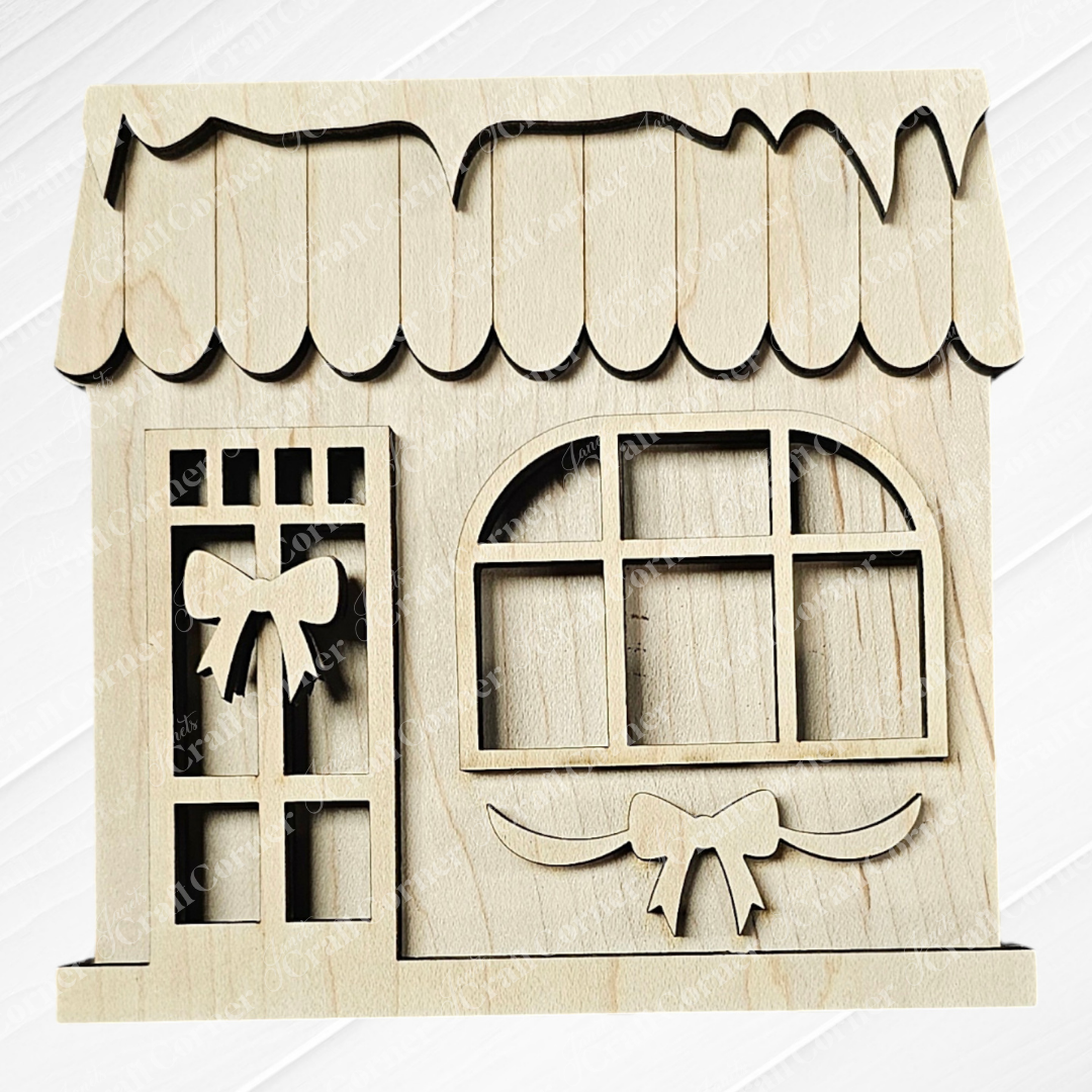 Discover the Wooden cutout from Janet's Craft Corner, featuring a charming small shop with an arched window, intricate door, and awning. Adorned with festive bows on the door and beneath the window, it serves as a delightful addition to your Christmas Village Shelf Sitters - Set of 4 | Early Access or as part of a DIY Craft Kit. The piece stands out beautifully against a subtle textured backdrop.