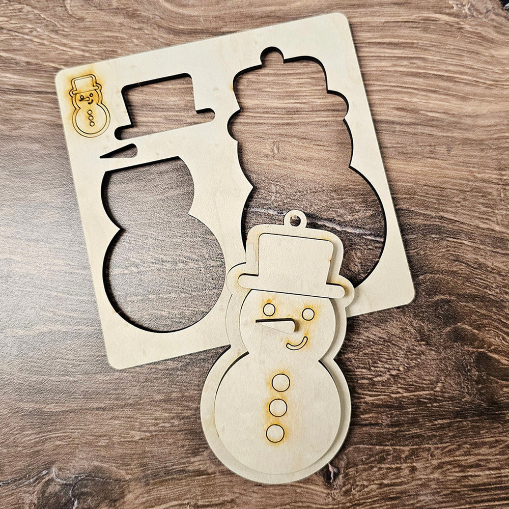 A wooden cutout showcasing a snowman shape with a top hat and scarf sits on a wooden table, accompanied by the stencil used to craft its snowman outline. Ideal for enthusiasts of Personalized Home Décor or those looking to add charm to DIY Craft Kit projects from Janet's Craft Corner, such as their "Sugar Cookie Ornaments - Set of 3.