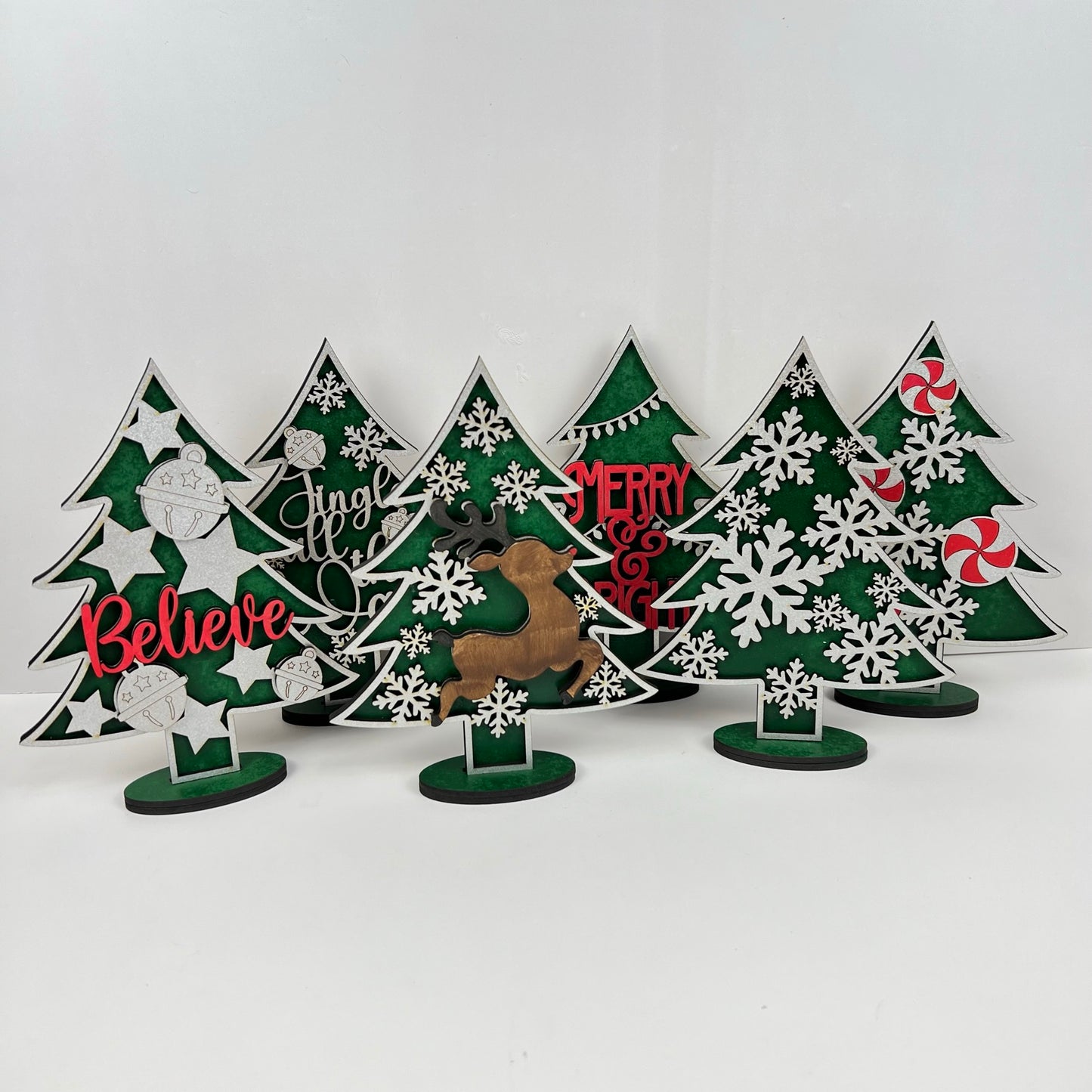 Janet's Craft Corner offers Standing Christmas Tree Shelf Sitters that are perfect for a shelf display. These decorative wooden trees feature words like "Believe," "Jingle," and "Merry" along with illustrations of snowflakes, ornaments, and candy. A reindeer silhouette adds to the festive holiday decor.