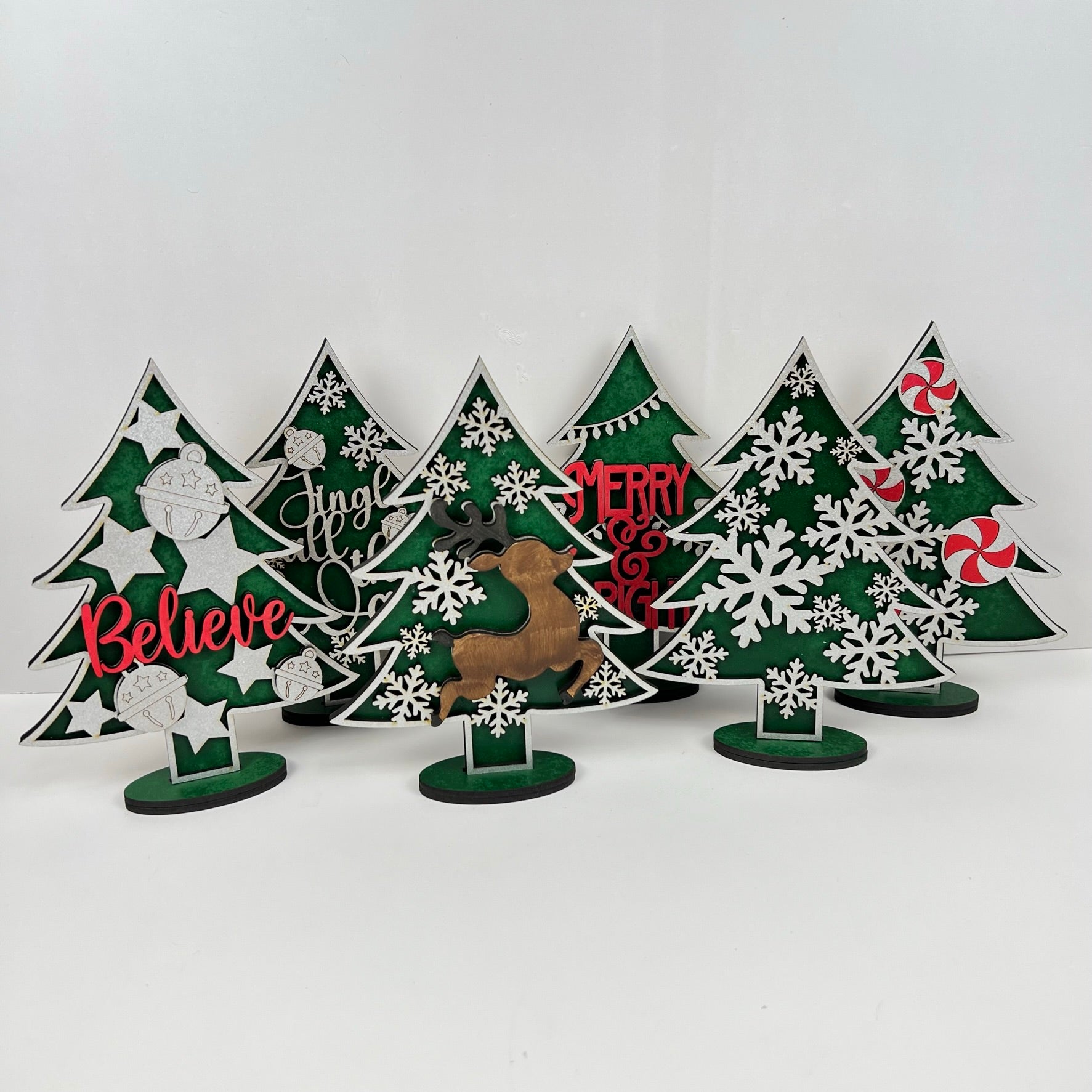 Janet's Craft Corner offers Standing Christmas Tree Shelf Sitters that are perfect for a shelf display. These decorative wooden trees feature words like "Believe," "Jingle," and "Merry" along with illustrations of snowflakes, ornaments, and candy. A reindeer silhouette adds to the festive holiday decor.