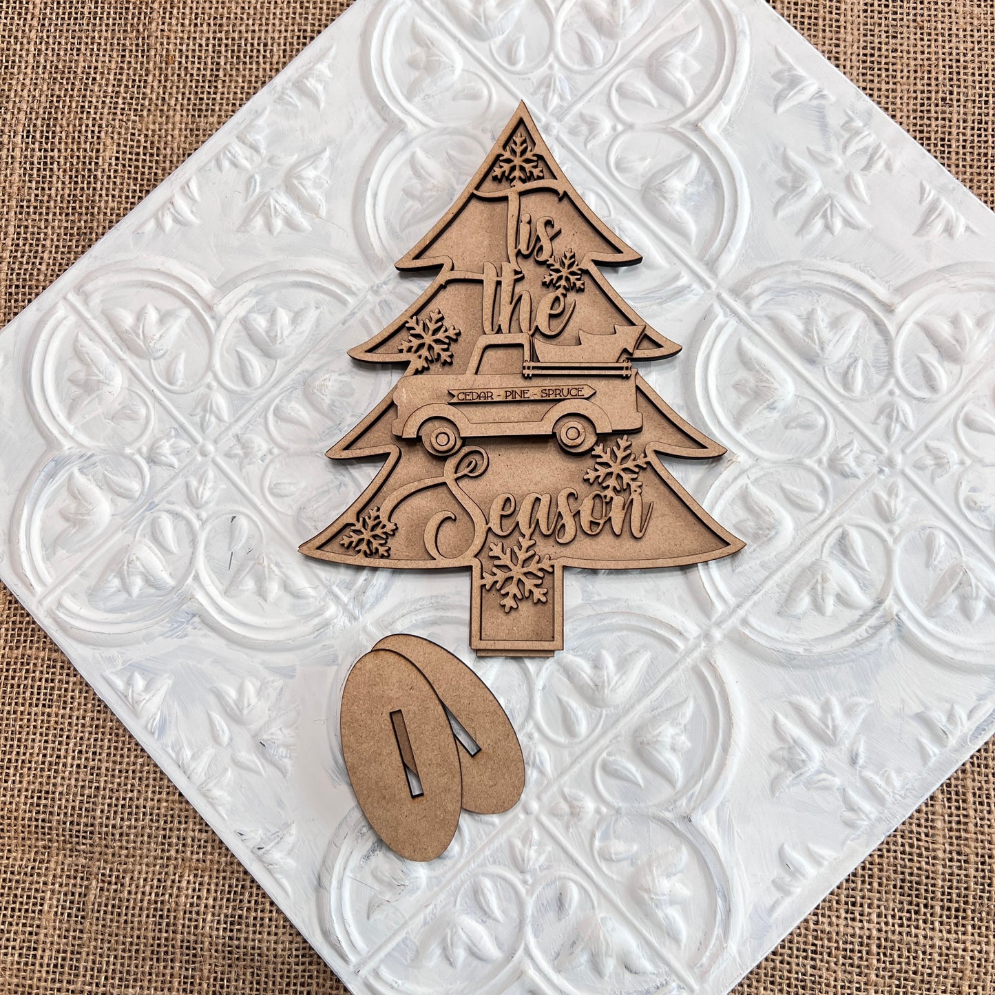 The Standing Christmas Tree Shelf Sitters from Janet's Craft Corner are beautifully designed wooden cutouts shaped like Christmas trees and adorned with "Tis the Season" text, a car, and snowflakes. These decorations are set on a decorative white tile with a burlap background, making them ideal holiday decor for any shelf display. The set comes with two wooden stand pieces for easy setup.
