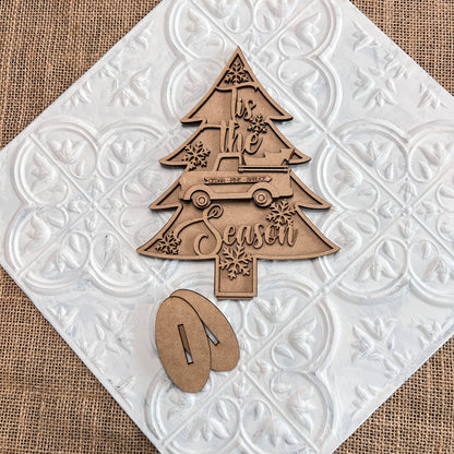 The Standing Christmas Tree Shelf Sitters from Janet's Craft Corner are beautifully designed wooden cutouts shaped like Christmas trees and adorned with "Tis the Season" text, a car, and snowflakes. These decorations are set on a decorative white tile with a burlap background, making them ideal holiday decor for any shelf display. The set comes with two wooden stand pieces for easy setup.