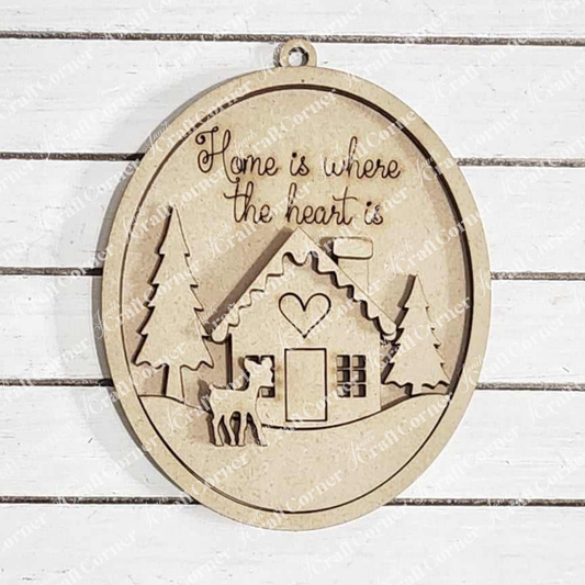 The Winter Cabin Ornament from Janet's Craft Corner is the perfect touch of personalized home décor, featuring an engraved house, heart, trees, and a moose. The text reads "Home is where the heart is," and it hangs elegantly on a white wooden wall.