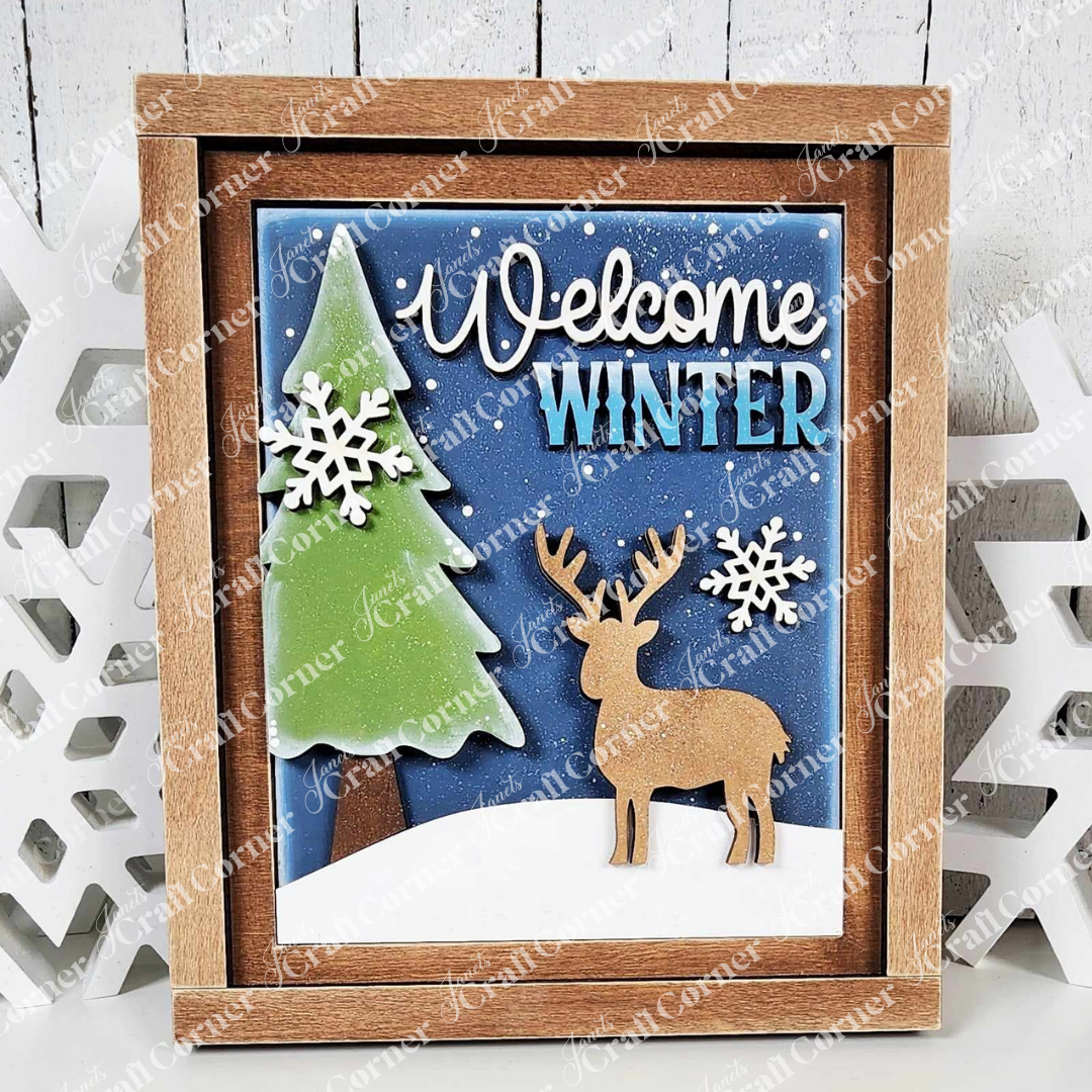 The "Welcome Winter Sign" by Janet's Craft Corner features a framed 3D winter scene with a pine tree, snowflakes, and a reindeer against a snowy blue backdrop. Its rustic wooden frame adds warmth to your home decor, inviting the season indoors with its welcoming message.