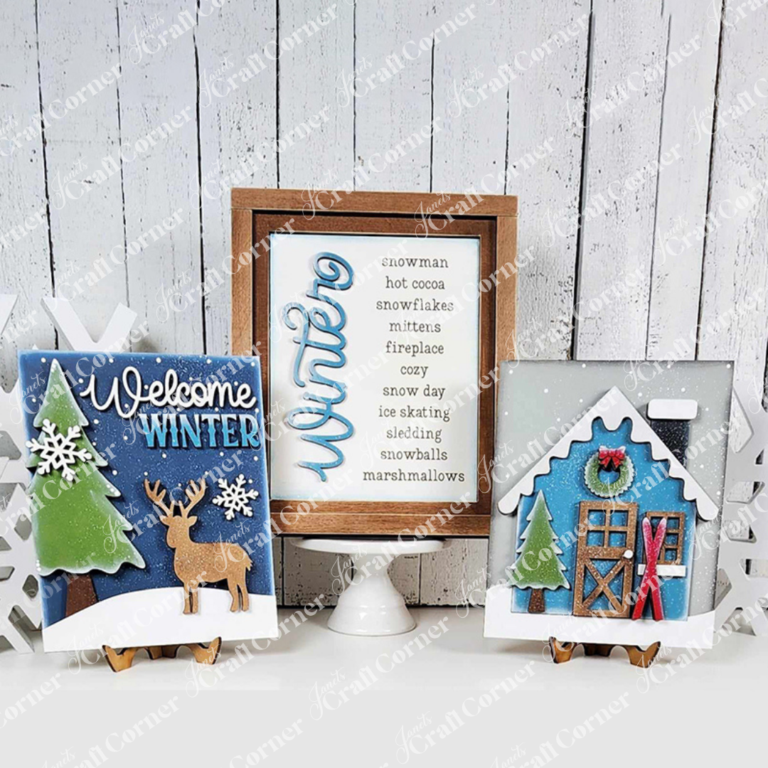 Nestled against a white wooden backdrop, the Winter Words Sign from Janet's Craft Corner brings a cozy vibe to your home decor. A framed list highlighting words of winter, accompanied by a "Welcome Winter" sign adorned with reindeer and snowflakes, along with a house embellished with festive greenery and snowy accents, offers perfect inspiration for your next DIY craft kit project.