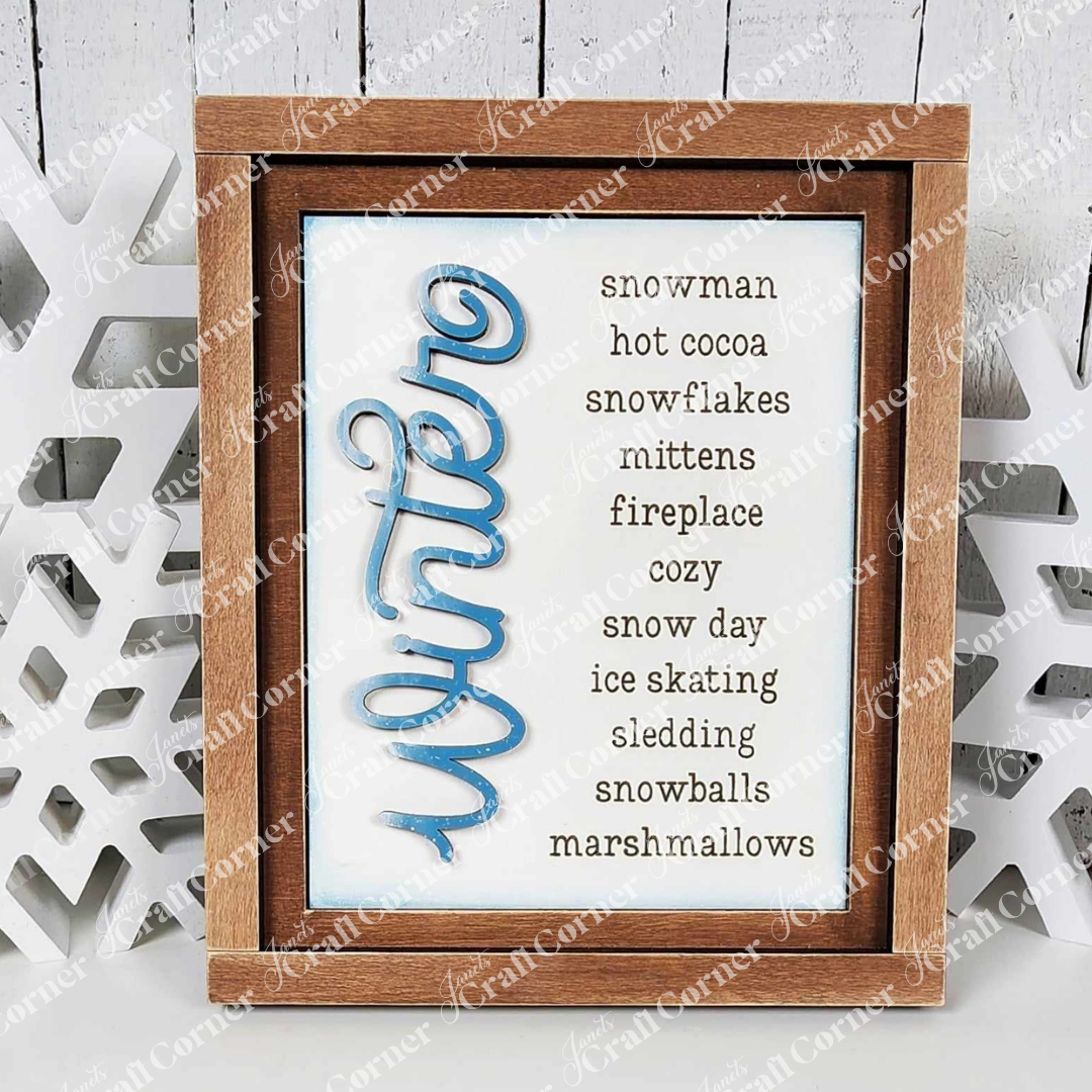 A framed "Winter Words Sign" from Janet's Craft Corner features elegant blue script on the left, with a charming list of words like snowman, hot cocoa, and snowflakes on the right. This piece is ideal for interchangeable home decor or as a DIY craft kit project. The background is beautifully accented by white snowflake decor.