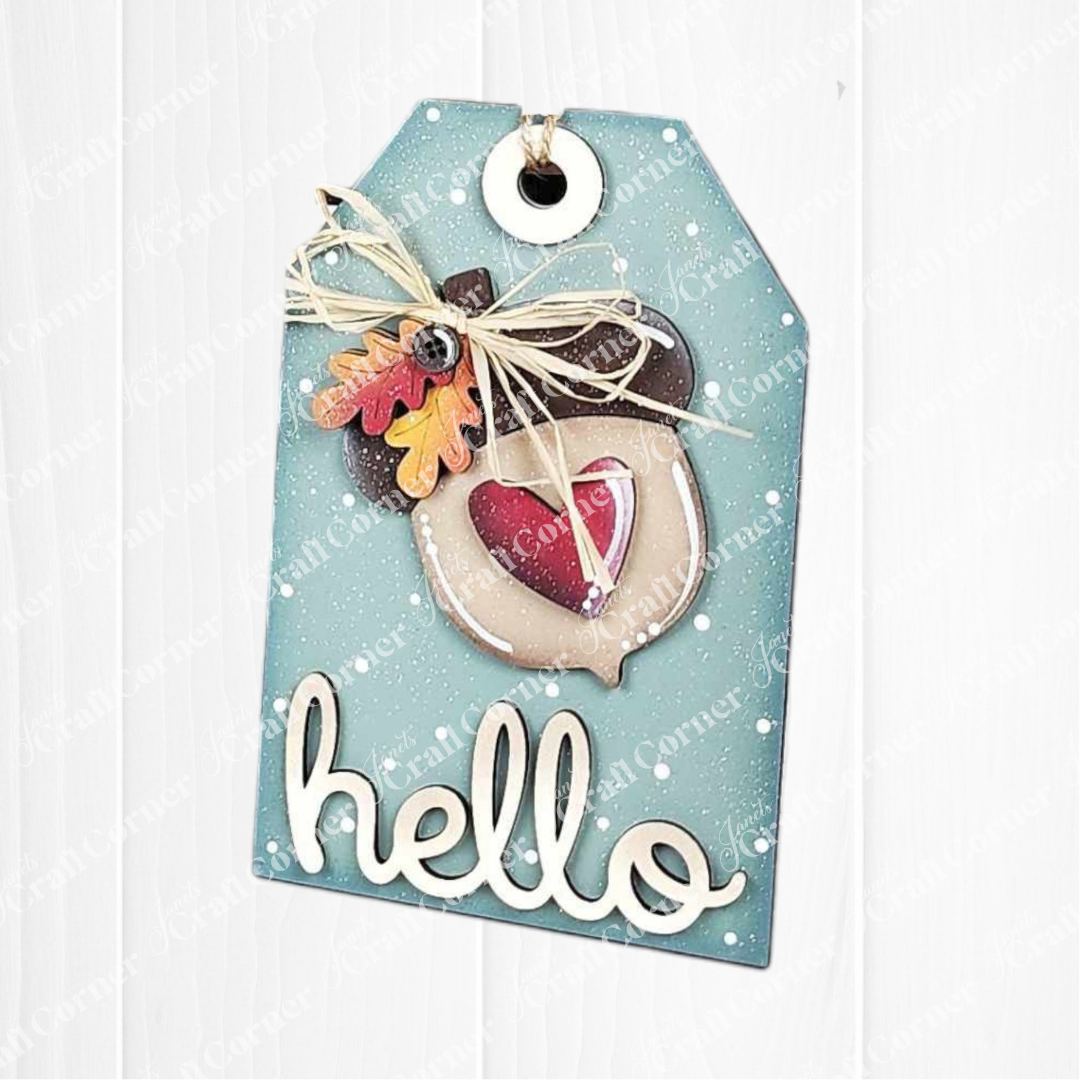 The Fall Harvest Mini Shelf Sitters by Janet's Craft Corner is a DIY home decor kit featuring a blue gift tag with twine bow, felt acorn with red heart, small leaves, and "hello"—perfect for autumn decorating or crafting.