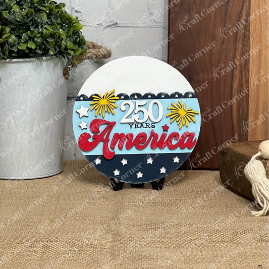 The America 250 Mini Shelf Sitters DIY craft kit by Janet's Craft Corner features a round patriotic sign with "250 Years America" in red, white, and blue, yellow fireworks, and stars—ideal for tiered tray displays or centerpieces.