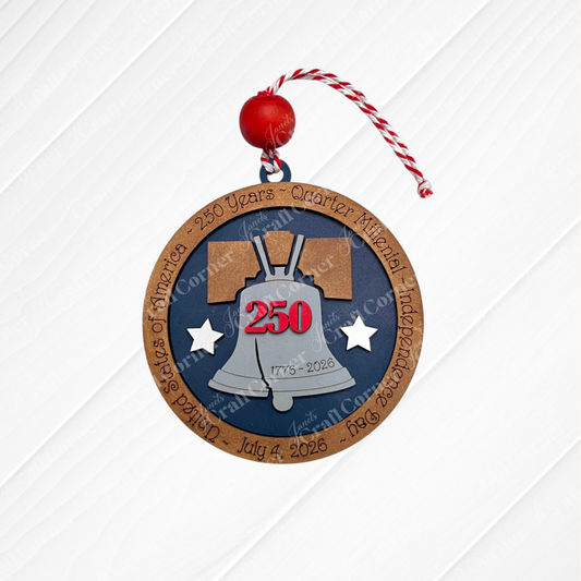 The Heartland America 250 Ornament - Painted is a round wood decoration with a red and white string, featuring a silver Liberty Bell, “250” in red, “1776-2026,” and the border reads “United States of America – 250 Years – Quarter America Independence Day – July 4, 2026.”.