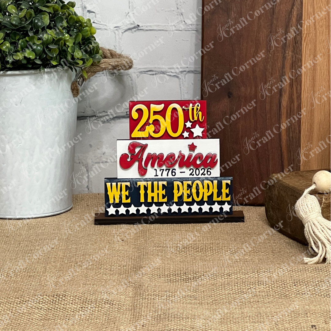 The America 250 Mini Shelf Sitters DIY craft kit by Janet's Craft Corner features a decorative block sign reading “250th America 1776-2026 WE THE PEOPLE” in red, yellow, and black—ideal for patriotic home décor displays.