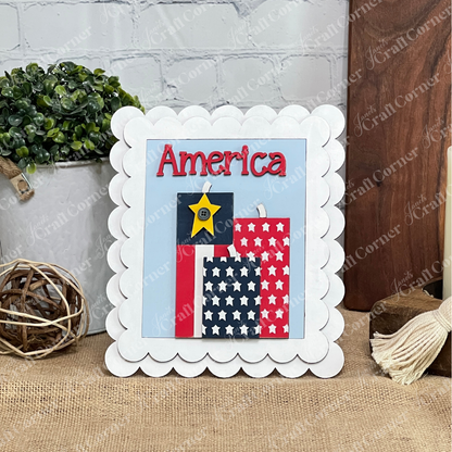 Janet's Craft Corner Patriotic Farmhouse Signs kit includes a framed sign with scalloped edges and “America” in red, above two flag designs and a yellow star—perfect tiered tray decor for your rustic DIY seasonal home.