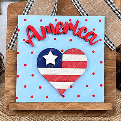 The Patriotic Squares DIY craft kit by Janet's Craft Corner features a decorative "America" sign with a heart-shaped USA flag on a blue, red polka-dot background—ideal for Independence Day décor or creative projects.