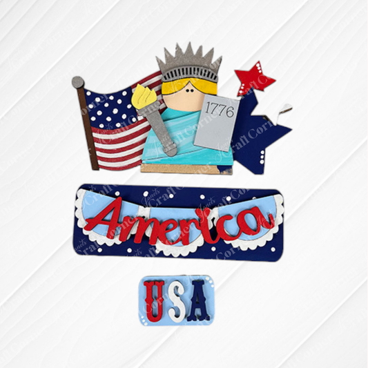 The America Interchangeable Insert DIY Craft Kit by Janet's Craft Corner features a cartoon Statue of Liberty, flag, and "America" and "USA" signs on a white wood background—ideal patriotic farmhouse décor for festive charm.