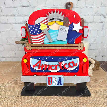 The America Interchangeable Insert DIY home decor kit by Janet's Craft Corner is a charming decorative red wooden truck adorned with a "USA" license plate and an "America" sign, embodying Americana spirit. It's filled with patriotic items such as a miniature Statue of Liberty, an American flag, and a star—ideal for crafting against the backdrop of a white brick wall.