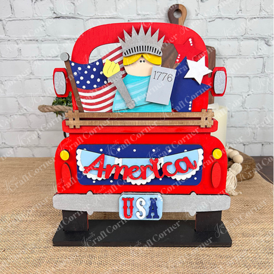 The America Interchangeable Insert DIY home decor kit by Janet's Craft Corner is a charming decorative red wooden truck adorned with a "USA" license plate and an "America" sign, embodying Americana spirit. It's filled with patriotic items such as a miniature Statue of Liberty, an American flag, and a star—ideal for crafting against the backdrop of a white brick wall.