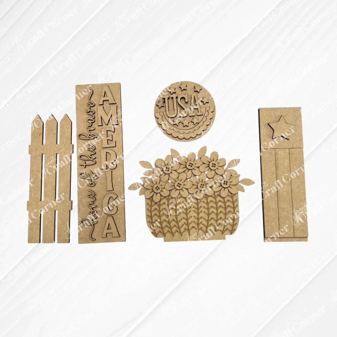 The America Interchangeable Porch Set by Janet's Craft Corner includes laser-cut wood pieces: a picket fence, two rectangular signs (one reads "Land of the free, America"), a USA round plaque, flower basket, and farmhouse accents board with star. Perfect for DIY décor!.