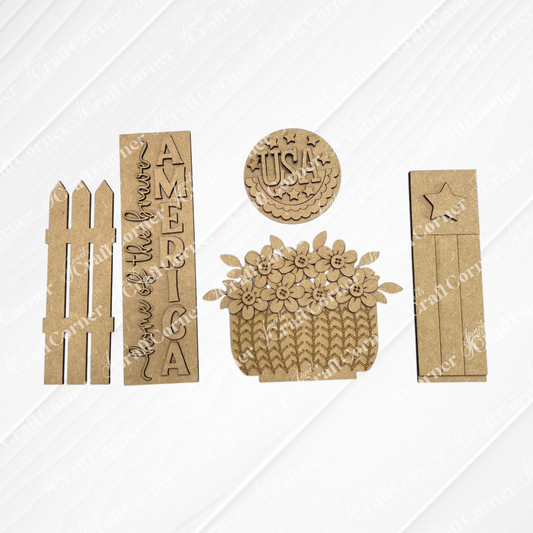 The America Interchangeable Porch Set by Janet's Craft Corner includes laser-cut wood pieces: a picket fence, two rectangular signs (one reads "Land of the free, America"), a USA round plaque, flower basket, and farmhouse accents board with star. Perfect for DIY décor!.