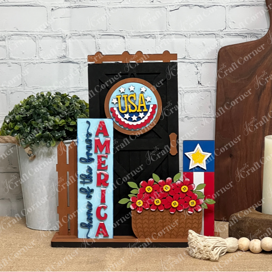 Janet's Craft Corner Farmhouse Porch Interchangeable Base is a DIY seasonal home decor kit featuring patriotic farmhouse accents—black door with USA sign, "America land of the free" sign, red flowers, and blue star block on a wood base.