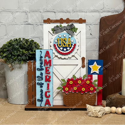 The Farmhouse Porch Interchangeable Base by Janet's Craft Corner is a DIY seasonal home decor craft kit featuring patriotic accents like a white barn door, "USA" sign, vertical "AMERICA" letters, and more—perfect for your porch or seasonal display.