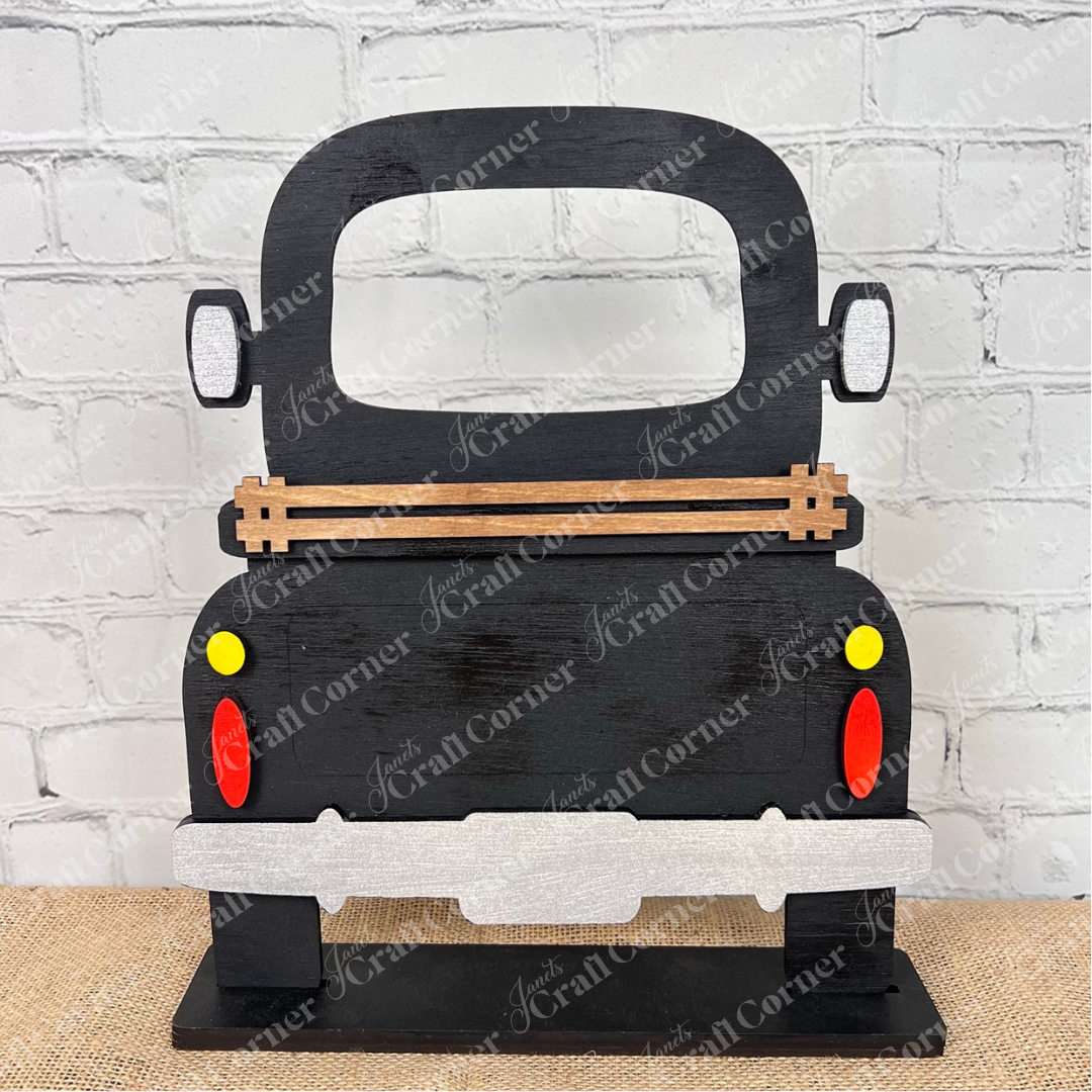 The Antique Truck Interchangeable Base  Standing DIY home decor kit by Janet's Craft Corner features wooden cutout pieces which have been painted black, displayed on a white brick background.