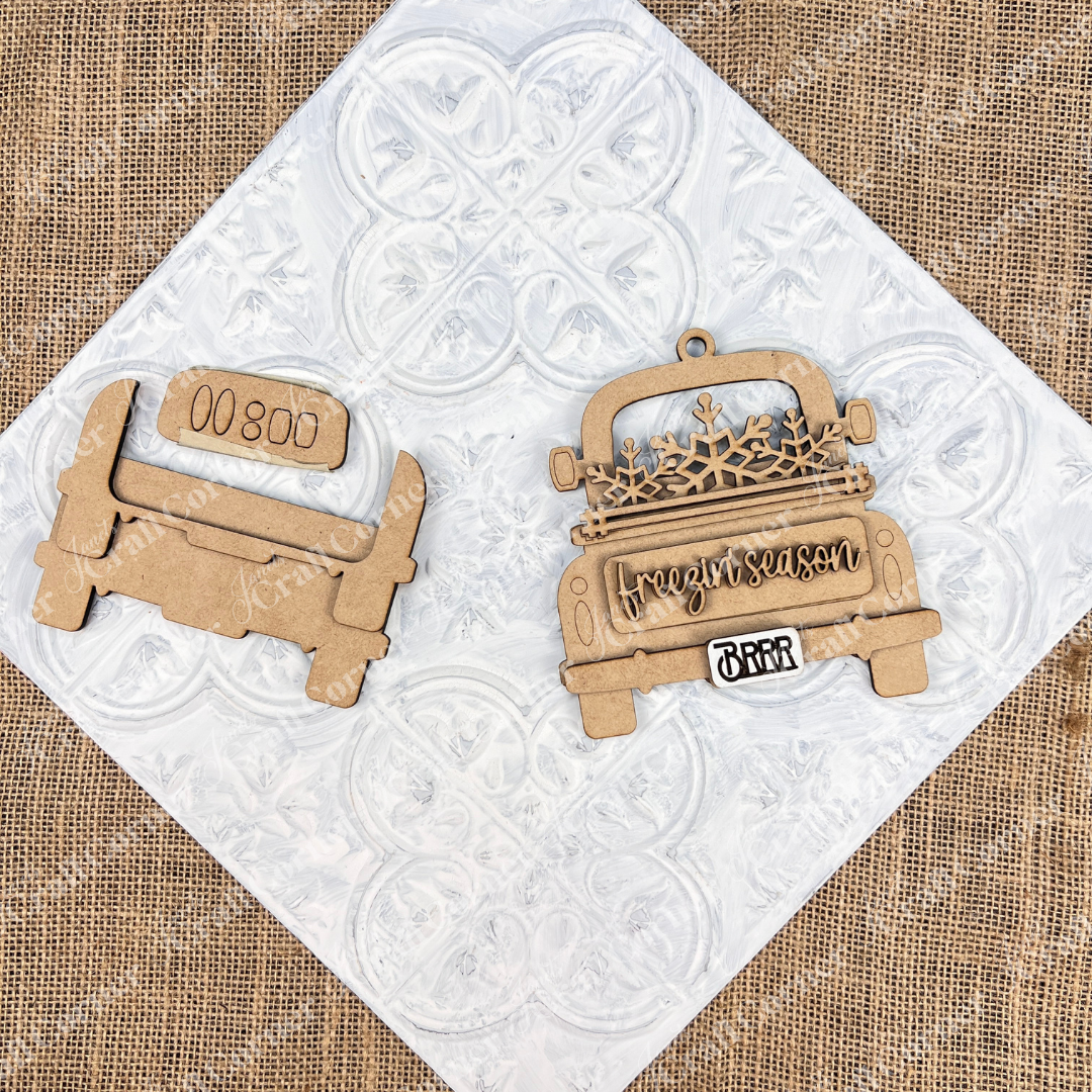 An antique truck ornament from Janet's Craft Corner's DIY Decoration Kits is placed on a textured white tile. The truck is adorned with "freezin' season" along with a snowflake design and the word "BRRR." Set against a rustic burlap background, this wooden piece is ideal for DIY Christmas décor.