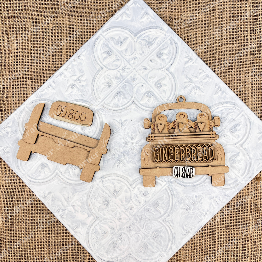 A wooden cutout depicting a vintage truck set against a white textured backdrop. An Antique Truck Ornament from Janet's Craft Corner's DIY Decoration Kits, showcases "Gingerbread Oh Snap" along with gingerbread figures and heart details on its bed. The scene rests atop patterned white tiles over burlap, making it ideal for Christmas décor.