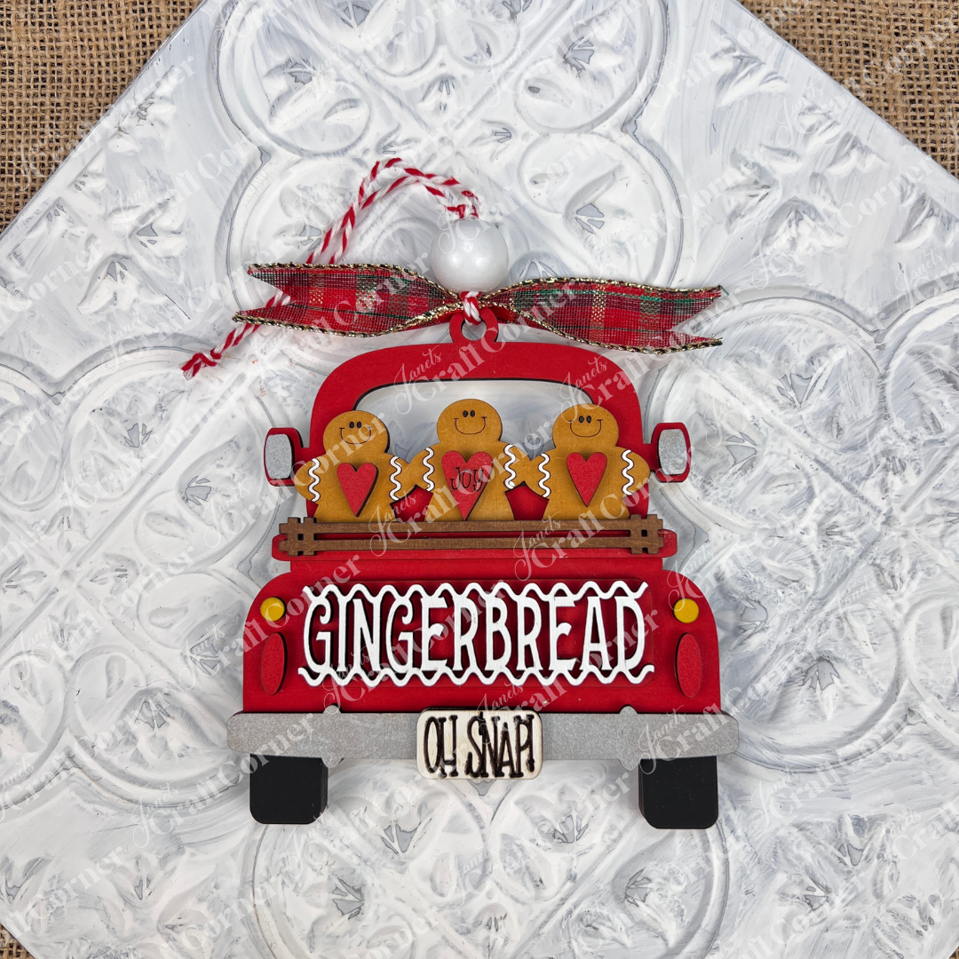 The Antique Truck Ornament from Janet's Craft Corner is a charming addition to your Christmas décor collection, featuring a red pickup loaded with gingerbread figures. The tailgate reads "GINGERBREAD" with "OH SNAP!" displayed below. A plaid bow atop the ornament completes the festive look, set against a textured white background.