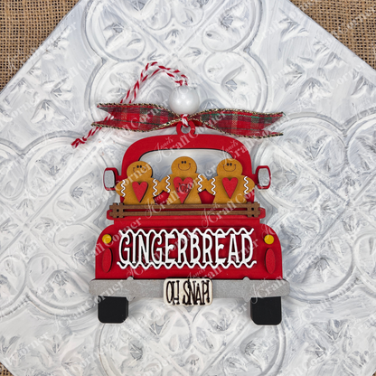 The Antique Truck Ornament from Janet's Craft Corner is a charming addition to your Christmas décor collection, featuring a red pickup loaded with gingerbread figures. The tailgate reads "GINGERBREAD" with "OH SNAP!" displayed below. A plaid bow atop the ornament completes the festive look, set against a textured white background.