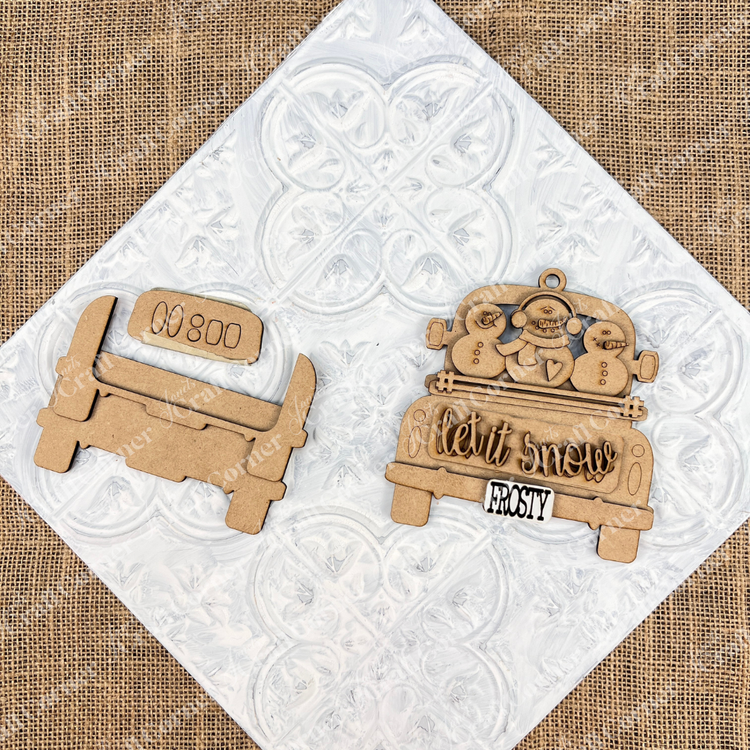 An antique truck ornaments from Janet's Craft Corner rest on a decorative white tile. It showcases "Let it Snow" with snowmen. This DIY decoration kits are perfect for Christmas décor, adding charm to any tree.