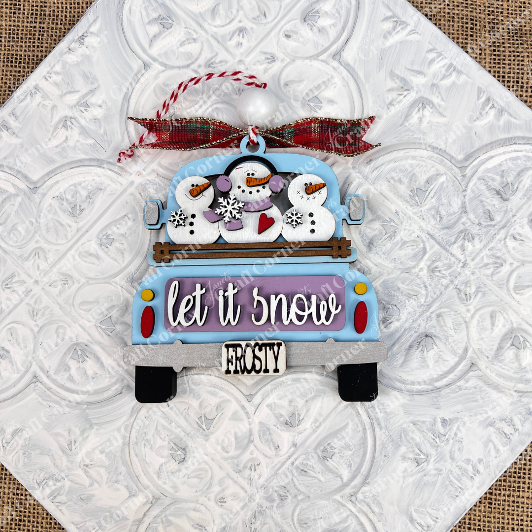 The "Antique Truck Ornament" from Janet's Craft Corner is a delightful addition to your Christmas décor, showcasing a charming blue antique truck with snowmen and a snowflake in the back, adorned with a "FROSTY" bumper sign. The text says "let it snow," beautifully accented by a red plaid ribbon against a textured white background.