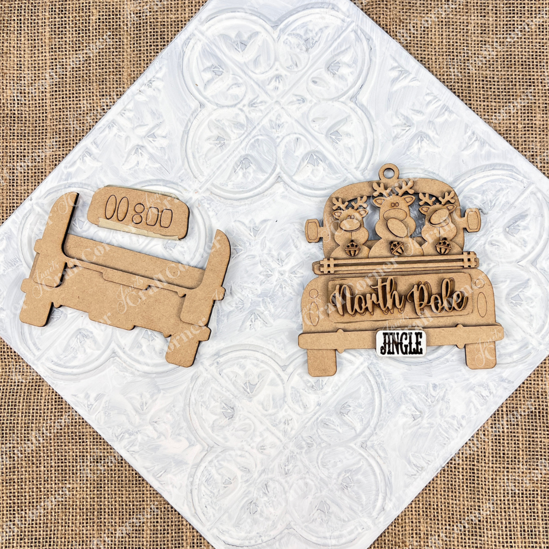 Displayed on a textured white and burlap background is a wooden cutout of a vintage truck. An Antique Truck Ornament from Janet's Craft Corner displays "North Pole," adorned with reindeer and a "Jingle" license plate. A perfect addition to your DIY decoration kits or Christmas décor collection.