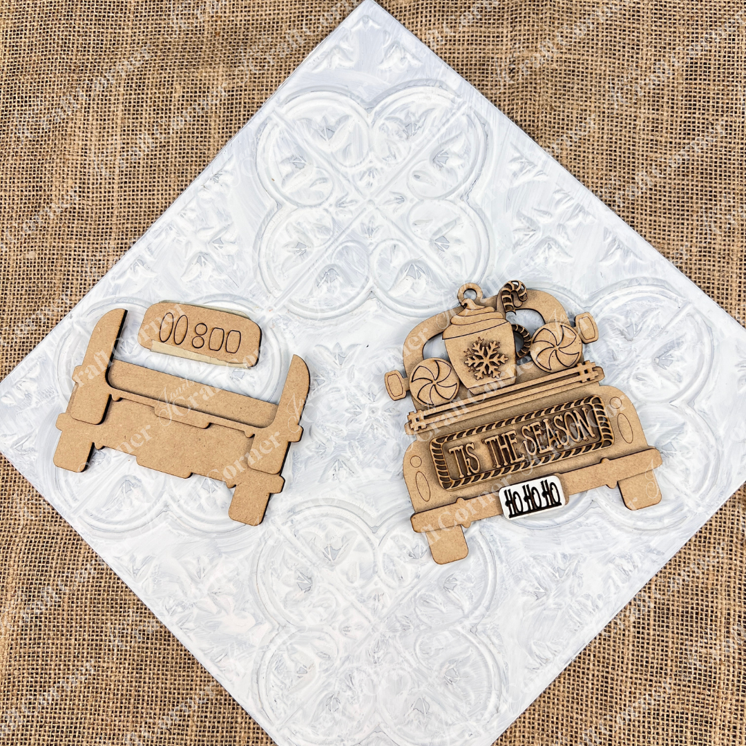On a textured white surface, a wooden craft ornament from Janet's Craft Corner are showcased as ideal additions to your Christmas décor. The ornament mimics the design of an antique truck with a festive messages such as "Tis the Season" and "Ho Ho Ho." The scene is set against a backdrop of burlap fabric.