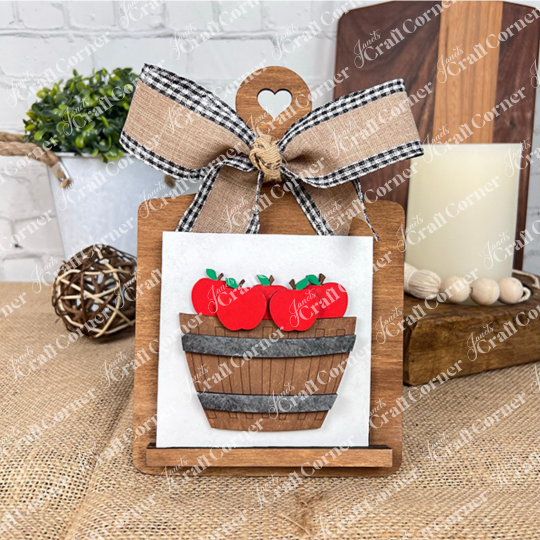 Janet's Craft Corner Apple Basket Square is displayed on a small wooden clipboard with a heart cutout and checkered bow, holding an apple-themed wood square—perfect DIY home decor for adding farmhouse charm to your rustic space.