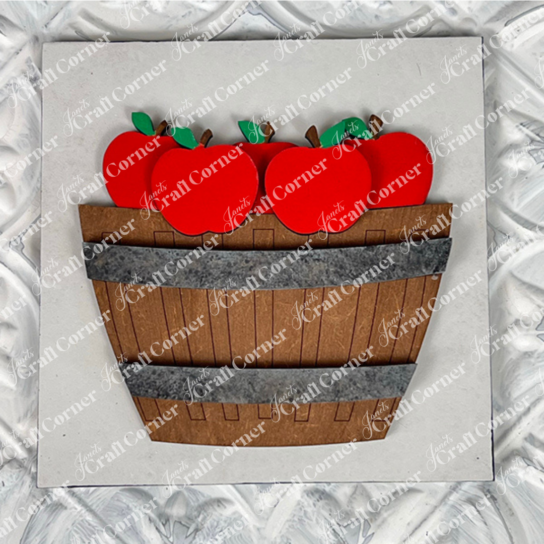 Janet's Craft Corner presents Apple Basket Square: a wood cut-outs craft kit featuring a wooden basket filled with bright red apples and green leaves, ideal for seasonal DIY decor, displayed on a textured white background.
