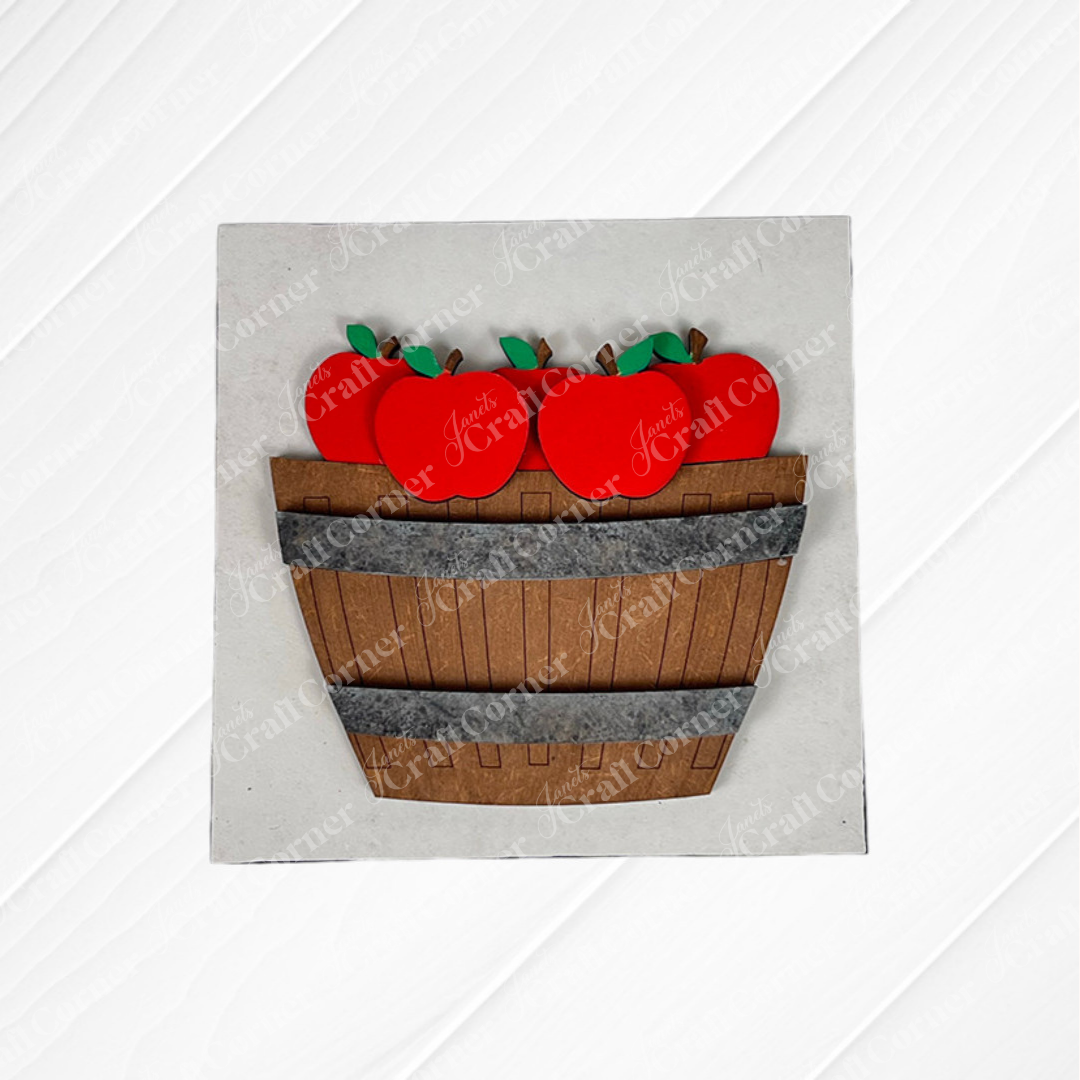 Janet's Craft Corner presents Apple Basket Square: a wood cut-outs craft kit featuring a wooden basket filled with bright red apples and green leaves, ideal for seasonal DIY decor, displayed on a textured white background.