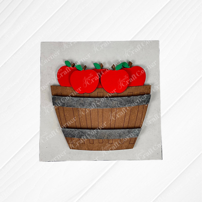 Janet's Craft Corner presents Apple Basket Square: a wood cut-outs craft kit featuring a wooden basket filled with bright red apples and green leaves, ideal for seasonal DIY decor, displayed on a textured white background.