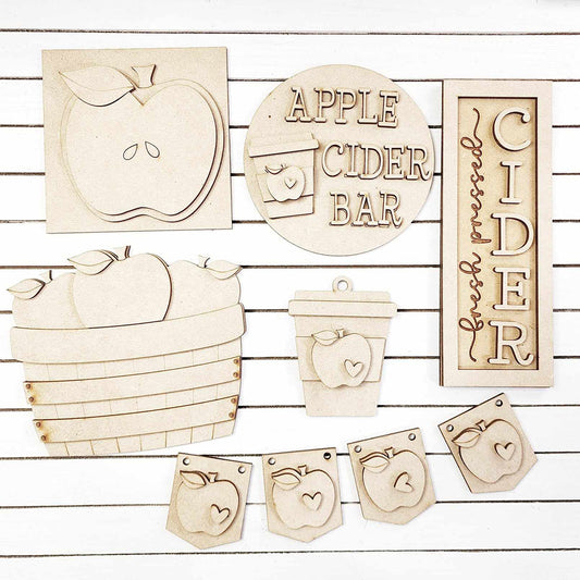 The Apple Cider Decor Set from Janet's Craft Corner features a charming collection of 6 wooden cutouts, each hand-painted with delightful apple-themed designs. This DIY home decor kit includes an apple shape, an "Apple Cider Bar" sign, a basket of apples, a coffee cup, and a banner adorned with apple icons. Perfect for craft enthusiasts looking to enhance their space with unique touches on a white striped background.