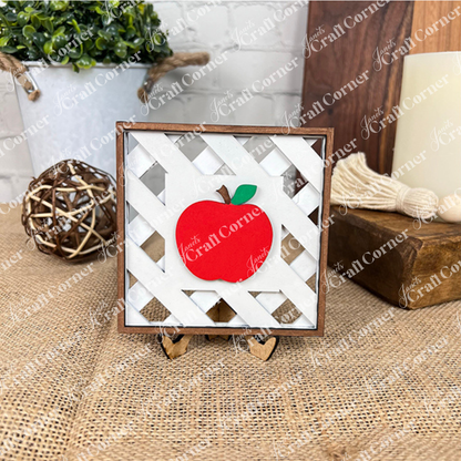 Janet's Craft Corner's Apple Swappable Squares DIY kit features a red apple wooden cut-out with a green leaf on a white lattice square frame and easel—ideal for rustic home decor and seasonal craft lovers. Set of 1.