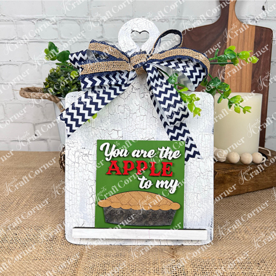Janet's Craft Corner presents the Apple Pie Square: a decorative white cutting board topped with chevron bow, greenery, and "You are the apple to my" pie sign—charming DIY decor for small spaces. Shown on a burlap table.