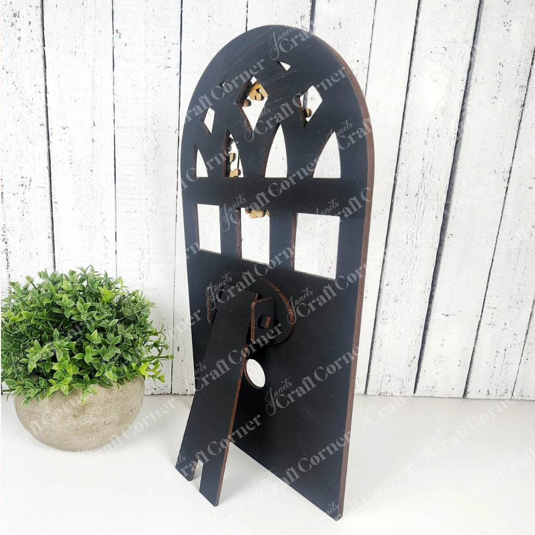 The Arch Window Interchangeable Frame for Squares by Janet's Craft Corner, shown in black beside a green potted plant and white plank wall, is the ideal DIY farmhouse décor kit.