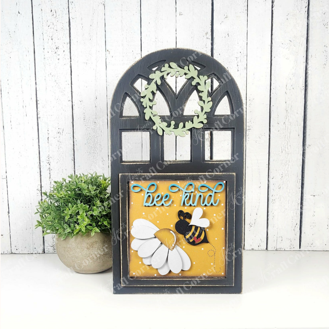 The Arch Window Interchangeable Frame for Squares by Janet's Craft Corner is a DIY farmhouse decor kit, shown with "bee kind" in blue, a bee, white flower, and green leaves beside a potted plant on a white wood plank background.