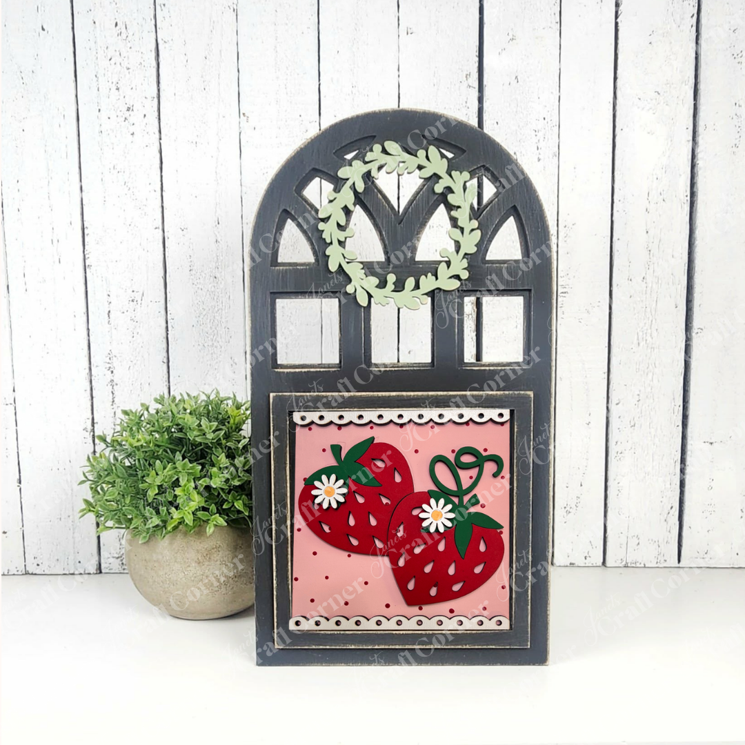 Janet's Craft Corner Arch Window Interchangeable Frame for Squares features a black frame, small green wreath, and pink strawberry panel—perfect as a DIY farmhouse décor kit, displayed with a potted plant on a white wooden background.