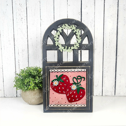 Janet's Craft Corner Arch Window Interchangeable Frame for Squares features a black frame, small green wreath, and pink strawberry panel—perfect as a DIY farmhouse décor kit, displayed with a potted plant on a white wooden background.