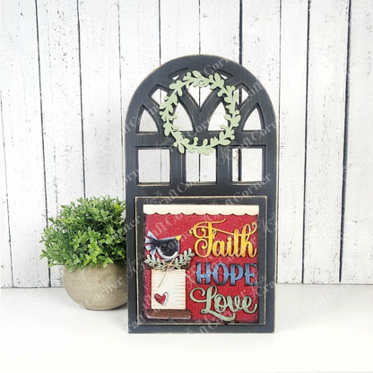 The Arch Window Interchangeable Frame for Squares by Janet's Craft Corner is a DIY farmhouse decor kit featuring a leafy wreath and "Faith Hope Love" plaque, shown beside a potted plant on a white wood background—perfect for charming home accents.