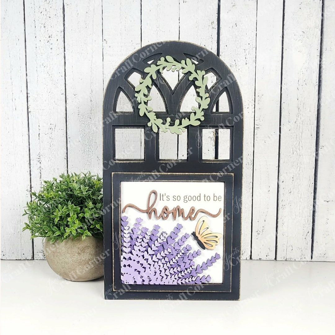 The Arch Window Interchangeable Frame for Squares by Janet's Craft Corner is a DIY farmhouse decor kit featuring a leafy wreath, purple lavender, and a yellow butterfly with "It's so good to be home" on a white wooden background.