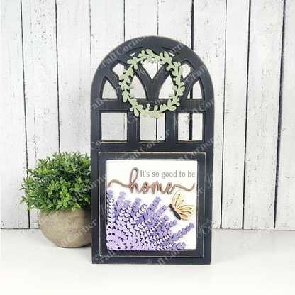 The Arch Window Interchangeable Frame for Squares by Janet's Craft Corner is a DIY farmhouse decor kit featuring a leafy wreath, purple lavender, and a yellow butterfly with "It's so good to be home" on a white wooden background.