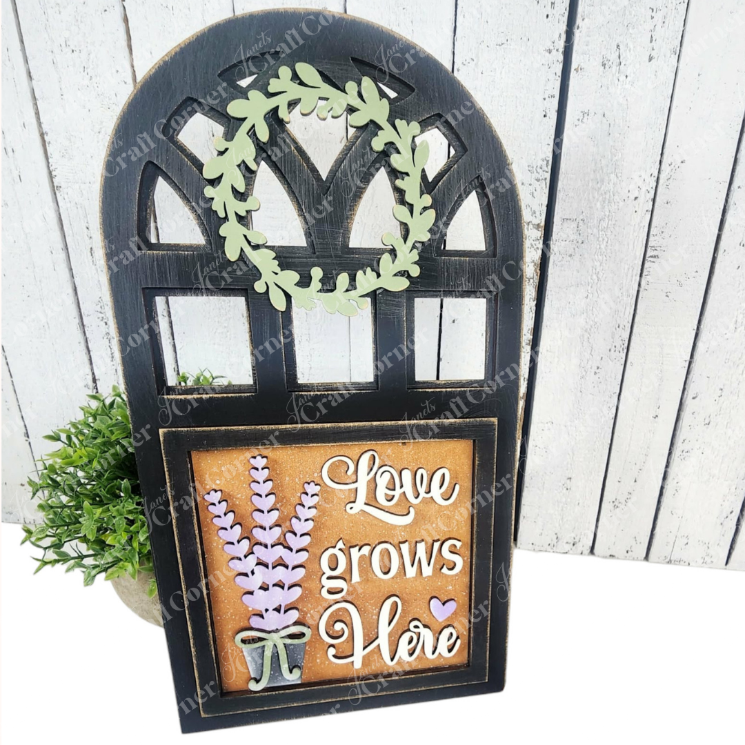 The Arch Window Interchangeable Frame for Squares by Janet's Craft Corner features a black frame, green leafy wreath, lavender cutouts, and "Love grows Here" in white—perfect DIY farmhouse décor with space for a small plant behind the sign.