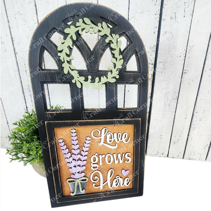The Arch Window Interchangeable Frame for Squares by Janet's Craft Corner features a black frame, green leafy wreath, lavender cutouts, and "Love grows Here" in white—perfect DIY farmhouse décor with space for a small plant behind the sign.